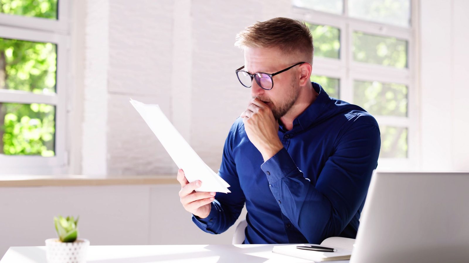 A man in glasses reading a printed report at a desk with his hand on his chin