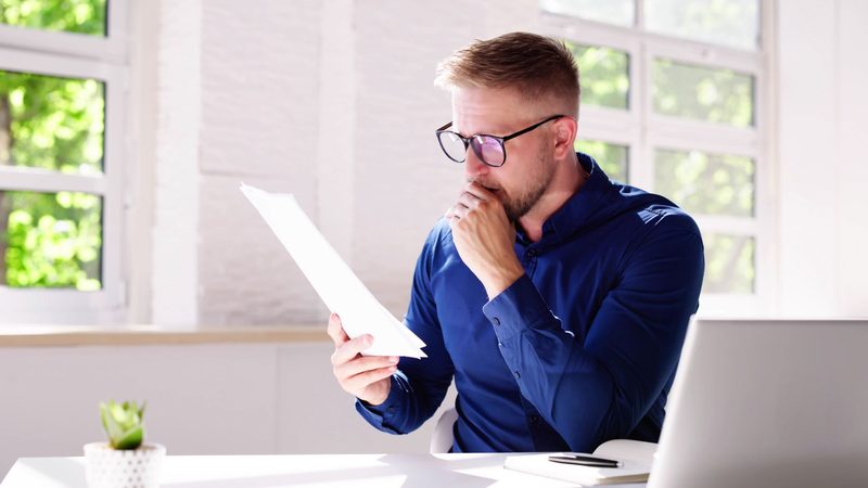 A man in glasses reading a printed report at a desk with his hand on his chin