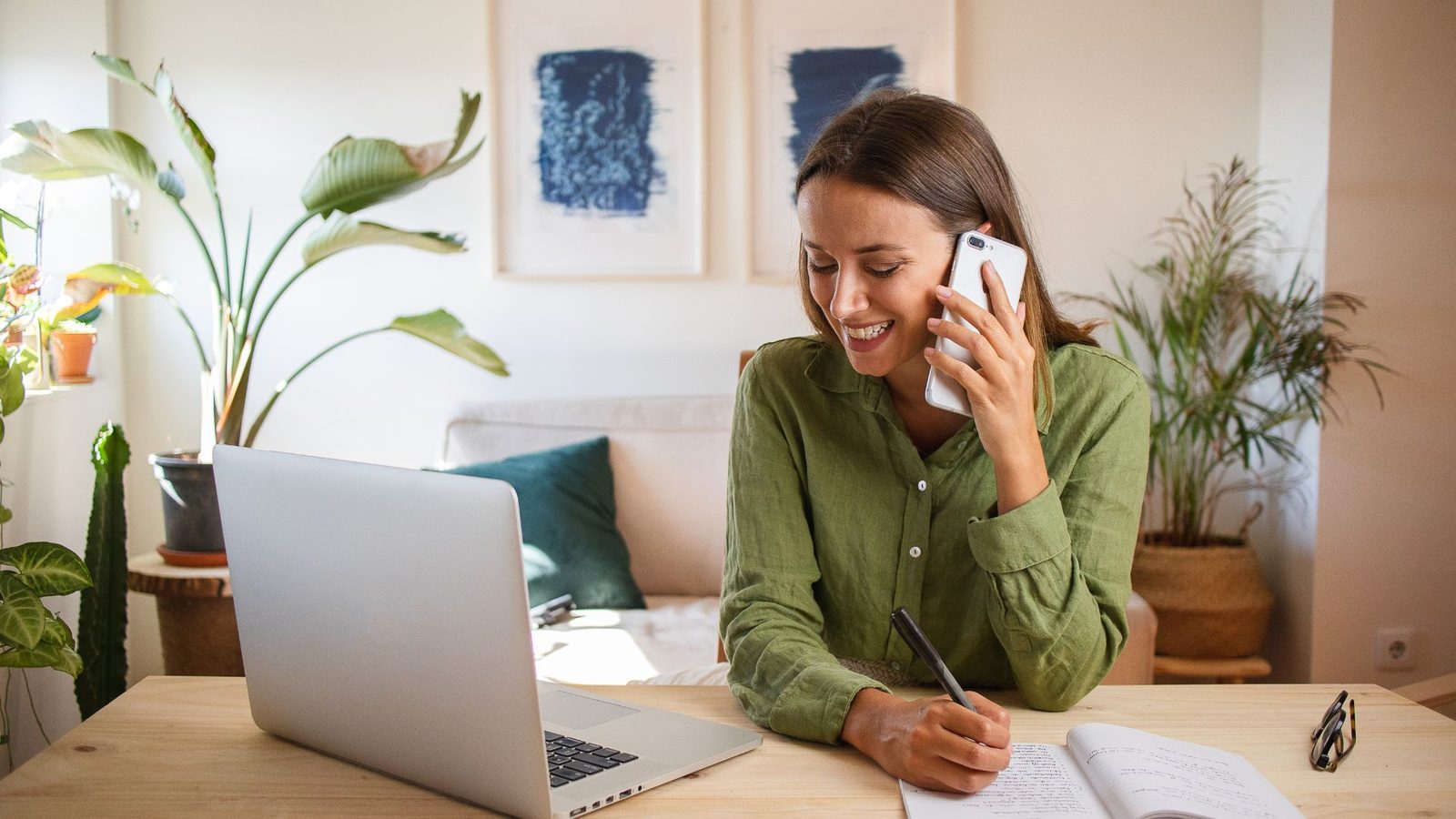 A woman on the phone taking notes at a table with a laptop open